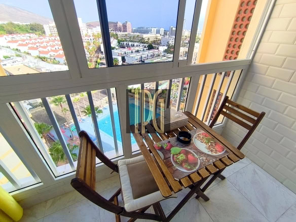 Balcony dining setup with a wooden table and two chairs, plates of food and a cup on placemats, overlooking a pool and cityscape beyond.