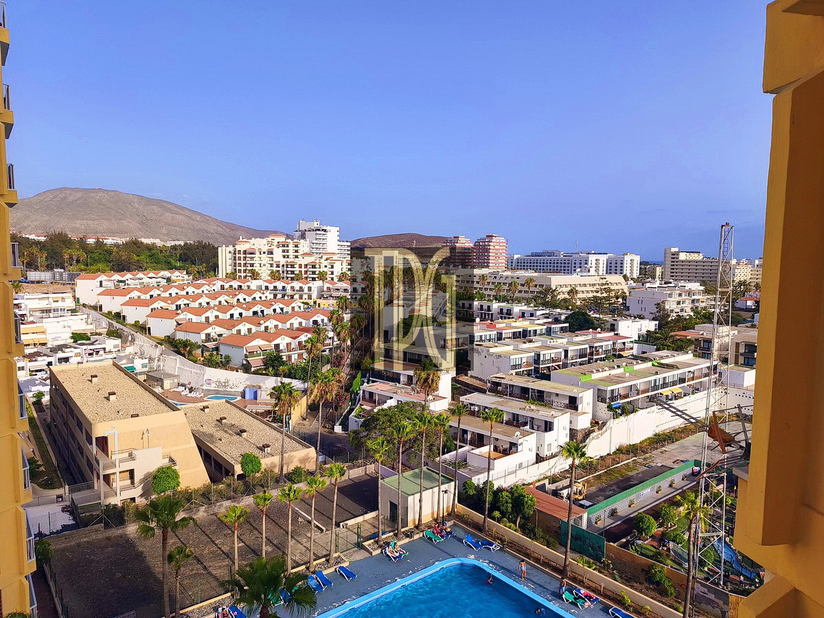 A sunny coastal city viewed from a high balcony: pool in the foreground, palm trees, white houses with red roofs, mid-rise buildings, and a mountain in the distance.
