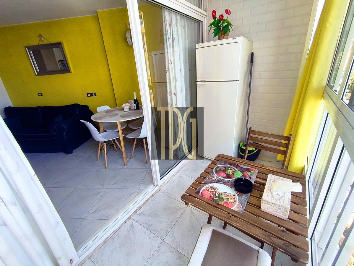 Balcony dining area with a wooden table and two chairs, a tissue box and placemats, and a white fridge; a bright yellow living room is visible through glass doors.