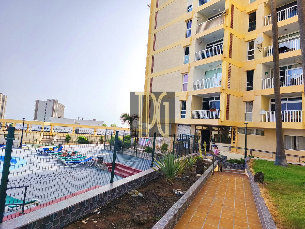 Yellow apartment building with open balconies overlooks a fenced pool deck and lounge chairs on a sunny day, with a person near the entrance on a tiled walkway.