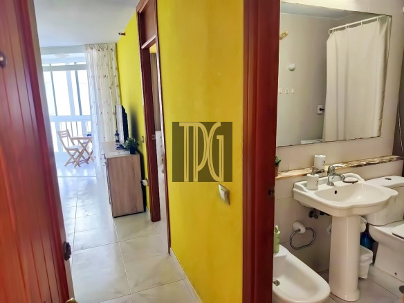 Hallway with bright yellow wall leading to a living area and balcony at left; bathroom doorway on the right with sink and mirror visible above a toilet and bidet area.