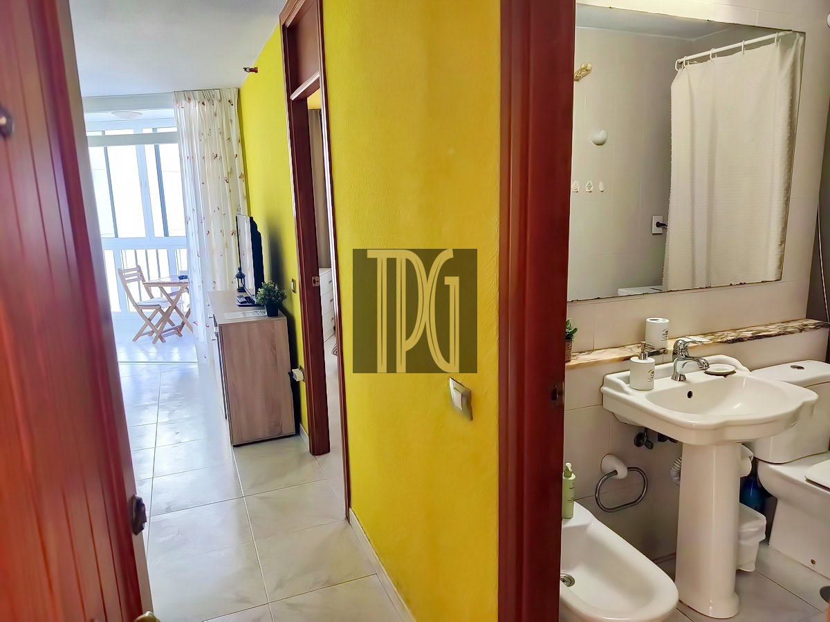 Hallway with bright yellow wall leading to a living area and balcony at left; bathroom doorway on the right with sink and mirror visible above a toilet and bidet area.