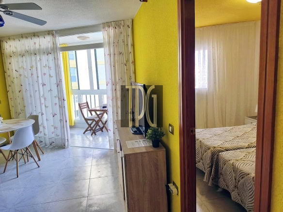 Bright yellow hotel studio with a round dining table and white chairs, TV on a wooden cabinet, and a balcony with a small table and chairs outside the sliding doors.