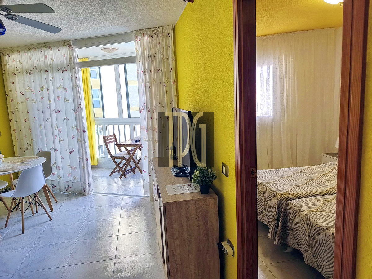 Bright yellow hotel studio with a round dining table and white chairs, TV on a wooden cabinet, and a balcony with a small table and chairs outside the sliding doors.