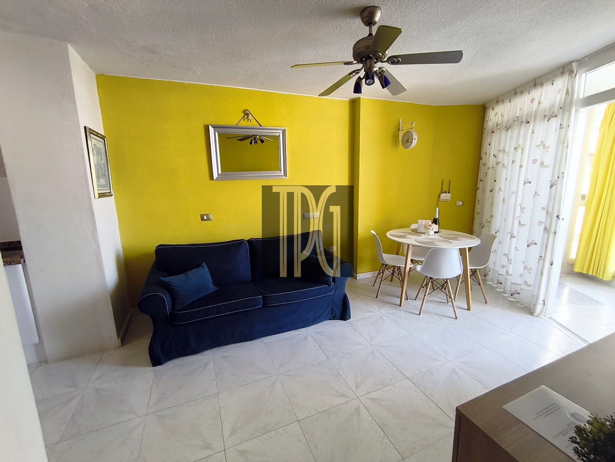 Bright yellow accent wall behind a navy blue sofa in a living-dining space, with a round white dining table and white chairs nearby.