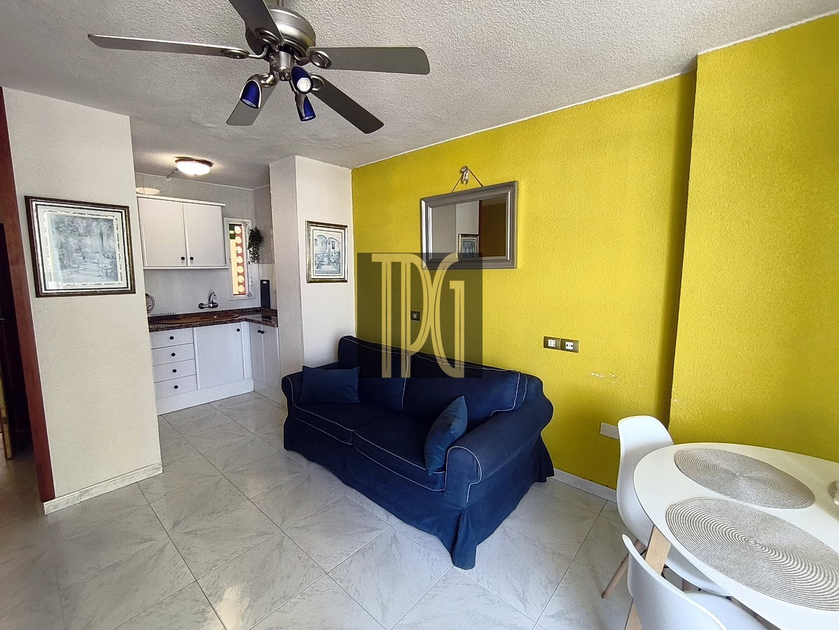 Open-concept living area with a navy sofa, round white dining table, and a bright yellow accent wall; small kitchen in the background.