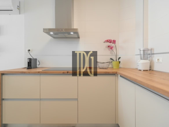 Modern L-shaped kitchen with beige cabinets, a wood countertop, stove and range hood, and a pink orchid near a blender.