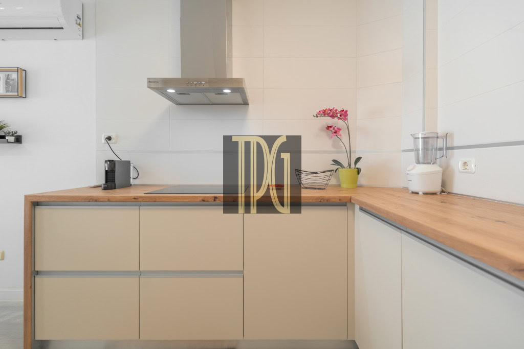 Modern L-shaped kitchen with beige cabinets, a wood countertop, stove and range hood, and a pink orchid near a blender.