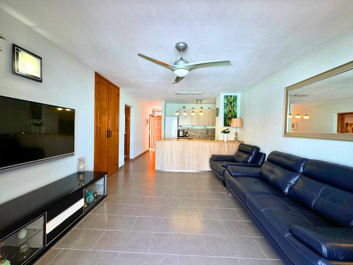 Open-concept living room with a black leather sectional, wall-mounted TV, and a kitchen bar in the background.