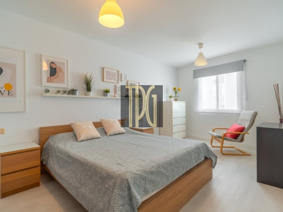 Bright modern bedroom with a wooden double bed, gray duvet, beige cushions, white nightstands, and a shelf of plants above the bed.