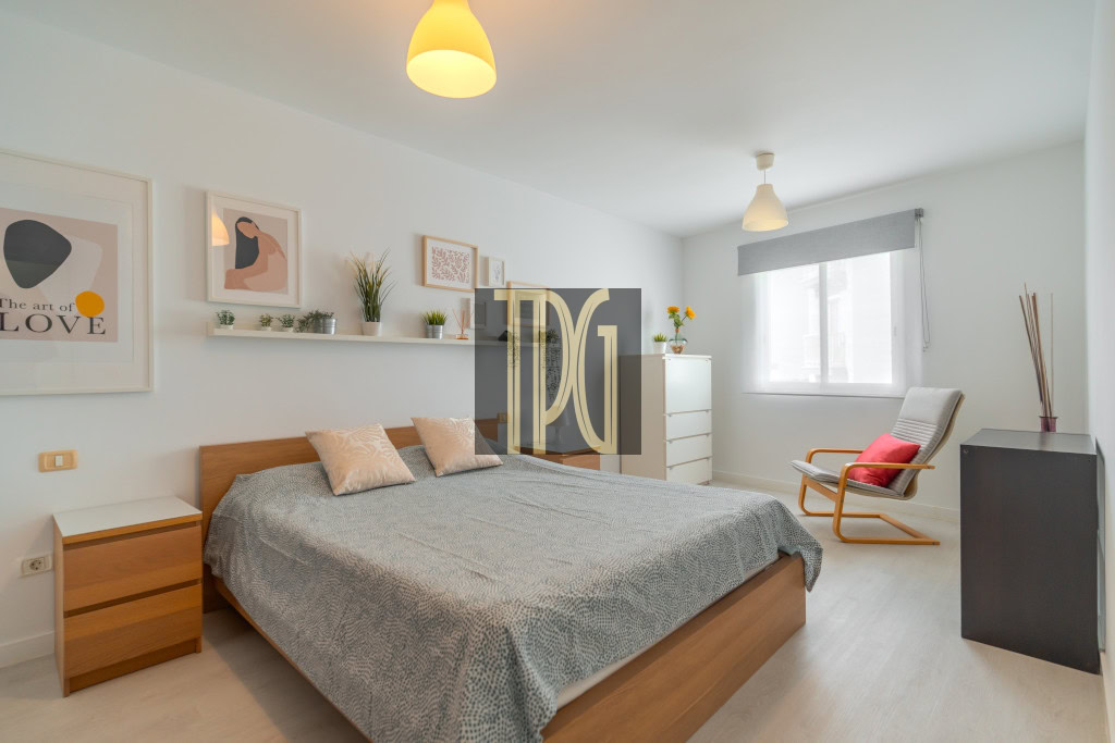 Bright modern bedroom with a wooden double bed, gray duvet, beige cushions, white nightstands, and a shelf of plants above the bed.