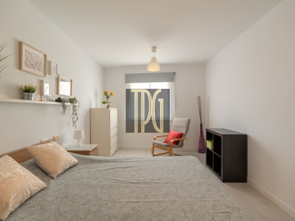 Light and airy minimalist bedroom with a low bed, gray patterned duvet, beige pillows, white walls, and a chair by the window.