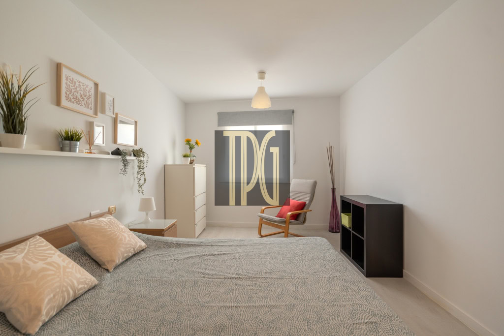 Light and airy minimalist bedroom with a low bed, gray patterned duvet, beige pillows, white walls, and a chair by the window.