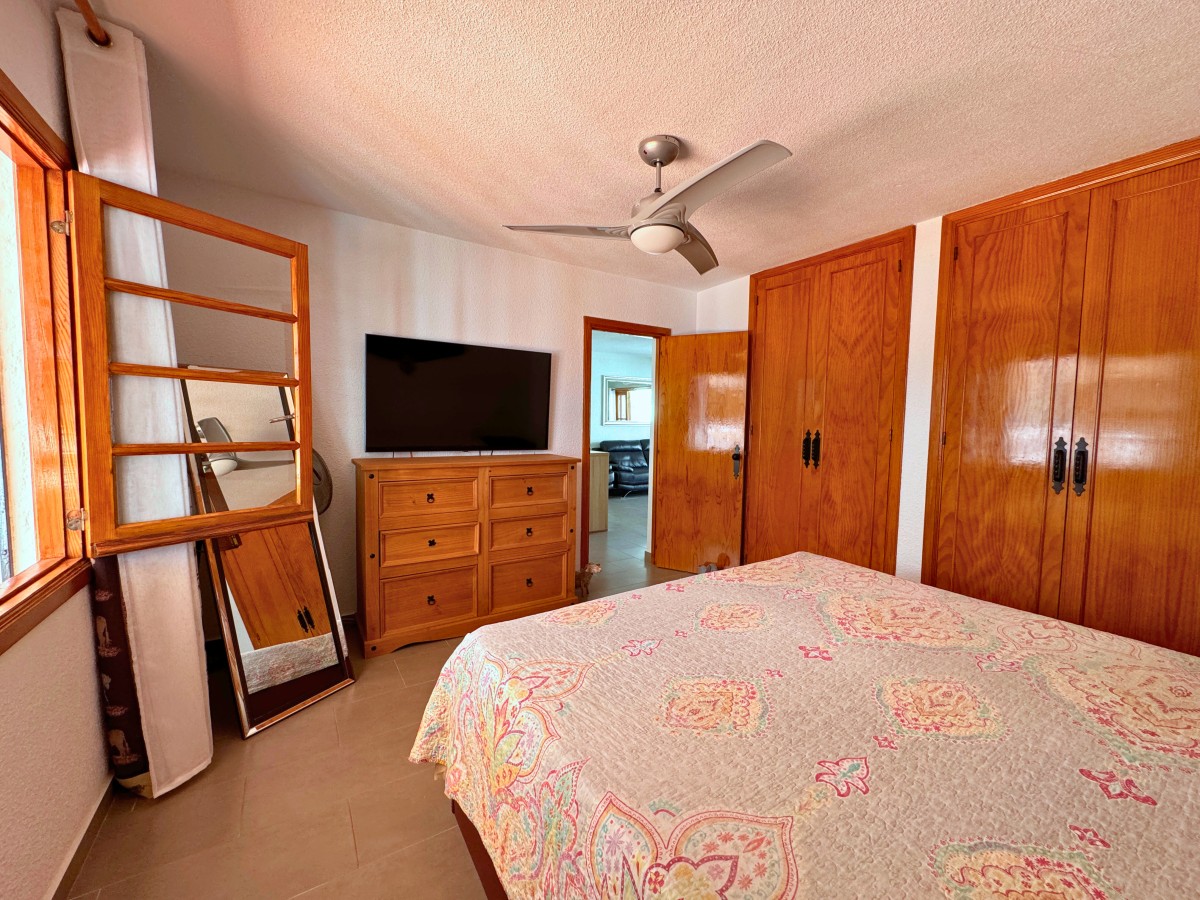 Bedroom with a quilted bedspread, wooden dresser with TV, and tall wooden closets; ceiling fan overhead and a mirror leaning by the window.