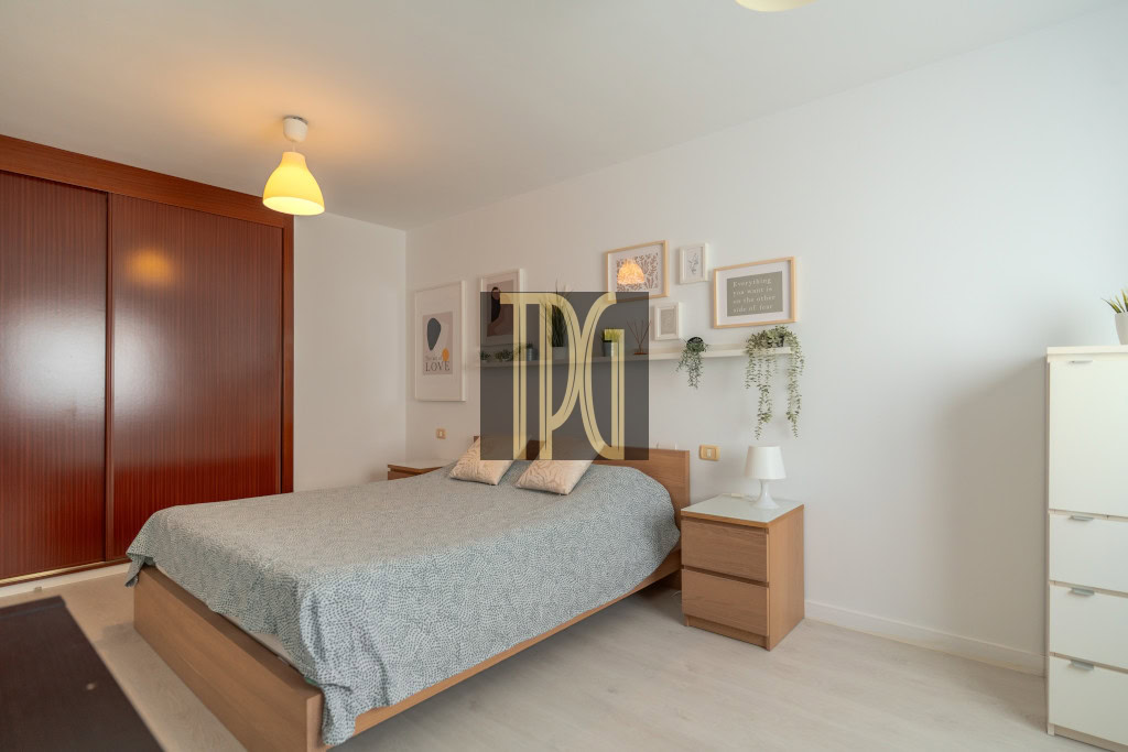 Cozy modern bedroom with a wooden bed frame, grey patterned duvet, two nightstands, and a gallery wall of framed art and plants.