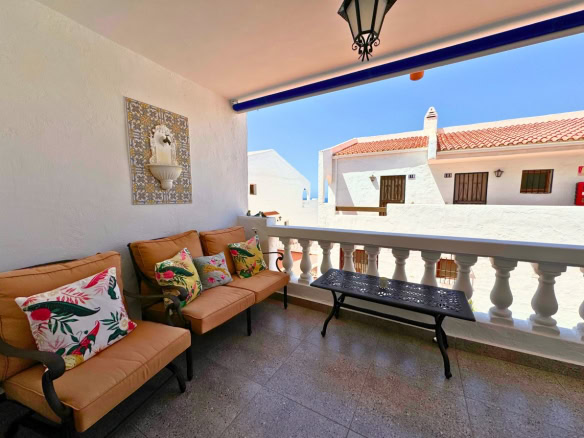 Outdoor balcony with orange cushioned seating and tropical-pattern pillows, a decorative metal table, and white balustrade under a blue sky.