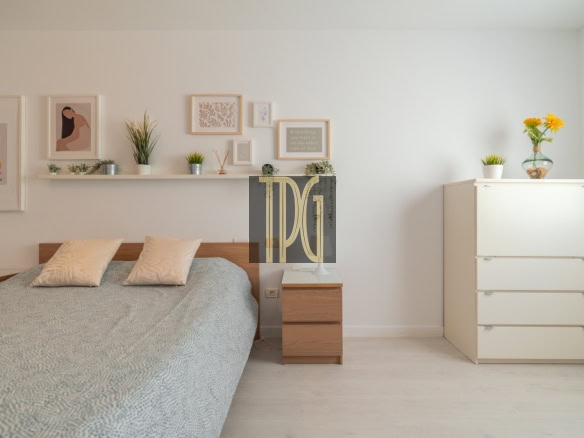 Modern bedroom with a low wooden bed, gray patterned duvet, and two pale cushions against a white wall gallery of framed art and a floating shelf with small plants.