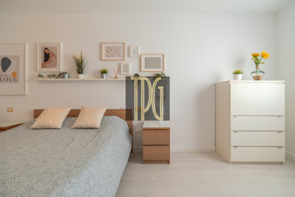 Modern bedroom with a low wooden bed, gray patterned duvet, and two pale cushions against a white wall gallery of framed art and a floating shelf with small plants.