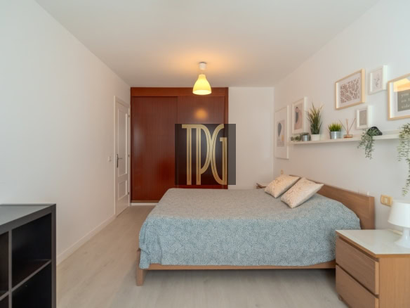 Bedroom with a wooden bed, blue-gray bedspread, and two matching pillows; white walls and a floating shelf with plants and framed art above the headboard