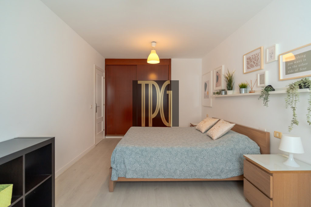 Bedroom with a wooden bed, blue-gray bedspread, and two matching pillows; white walls and a floating shelf with plants and framed art above the headboard