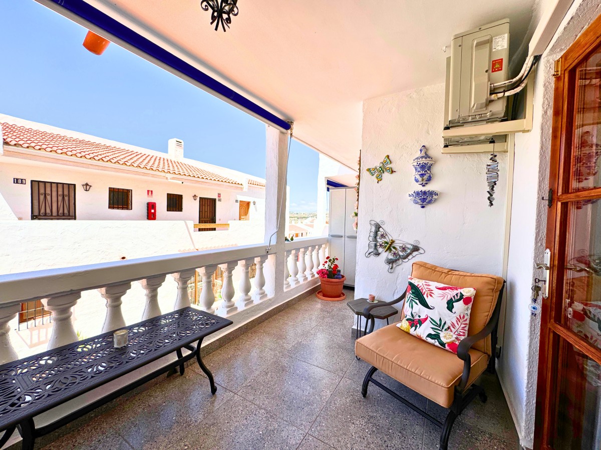 Cozy balcony seating area with a cushioned chair, floral pillow, and a black metal table, plus potted plant and blue-and-white ceramic wall decor on a white wall. Sunny view of neighboring white houses with tile roofs across the street.