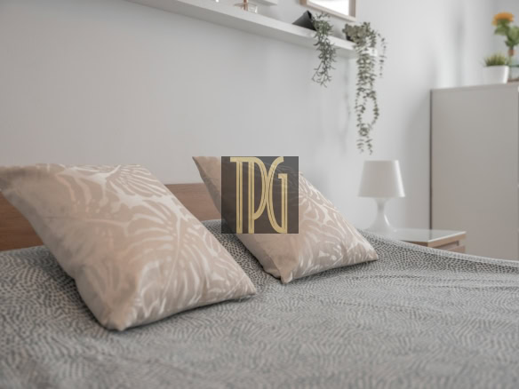 Bed with two beige patterned pillows and a gray patterned duvet in a bright, minimalist bedroom with a white cabinet and plants in the background.