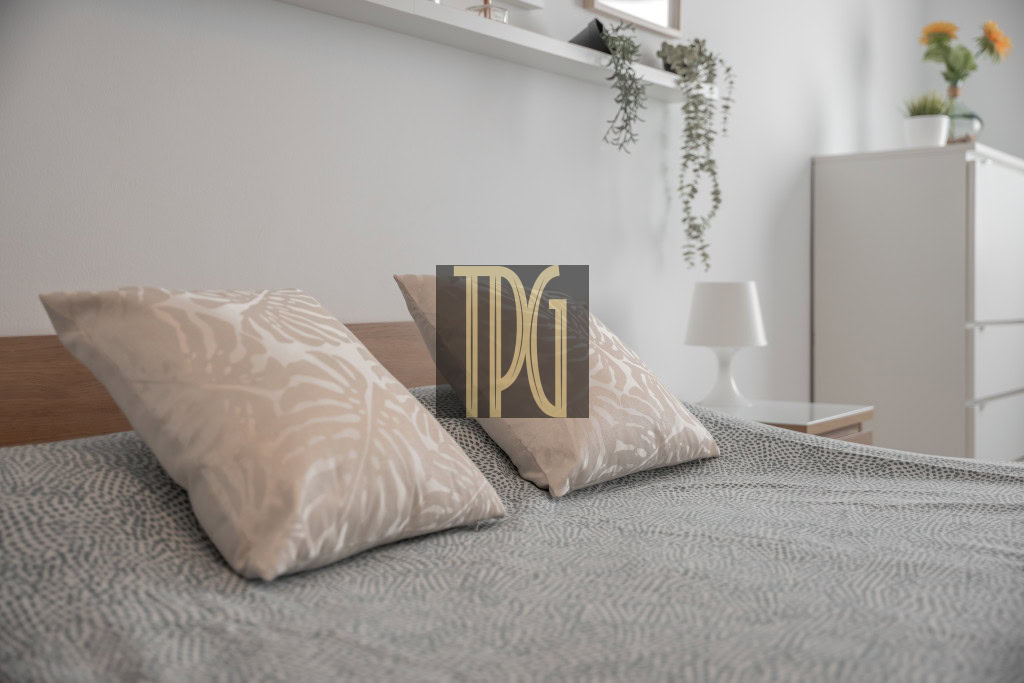 Bed with two beige patterned pillows and a gray patterned duvet in a bright, minimalist bedroom with a white cabinet and plants in the background.