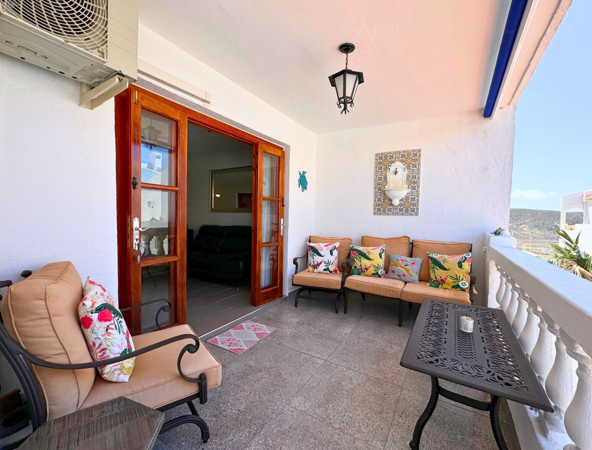 Sunny balcony lounge with beige cushioned seating and colorful tropical pillows, wooden French doors open to the living room.