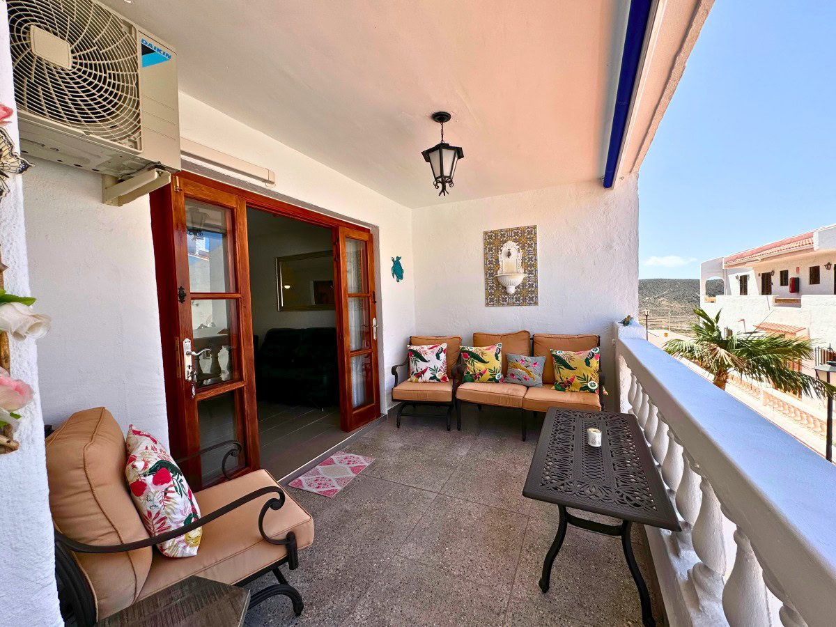Outdoor balcony seating area with beige cushioned chairs and tropical-pattern pillows, white walls, and a black ornate metal table. A glass door opens to an indoorsy living space; an air conditioner sits on the wall above. Sunny day glimpsed beyond a white railing with a view of neighboring buildings.