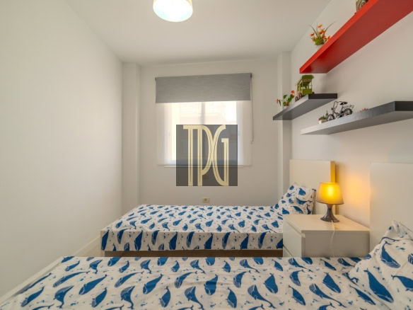 Twin bedroom with two single beds covered in blue shark-patterned bedding, white walls, and a nightstand with a yellow lamp between them