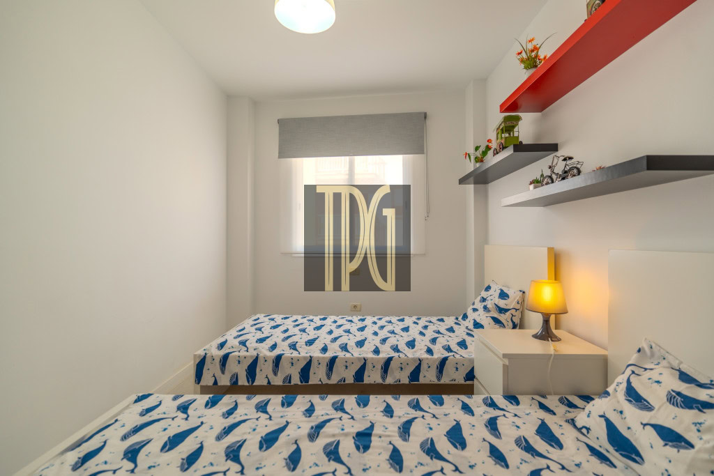 Twin bedroom with two single beds covered in blue shark-patterned bedding, white walls, and a nightstand with a yellow lamp between them