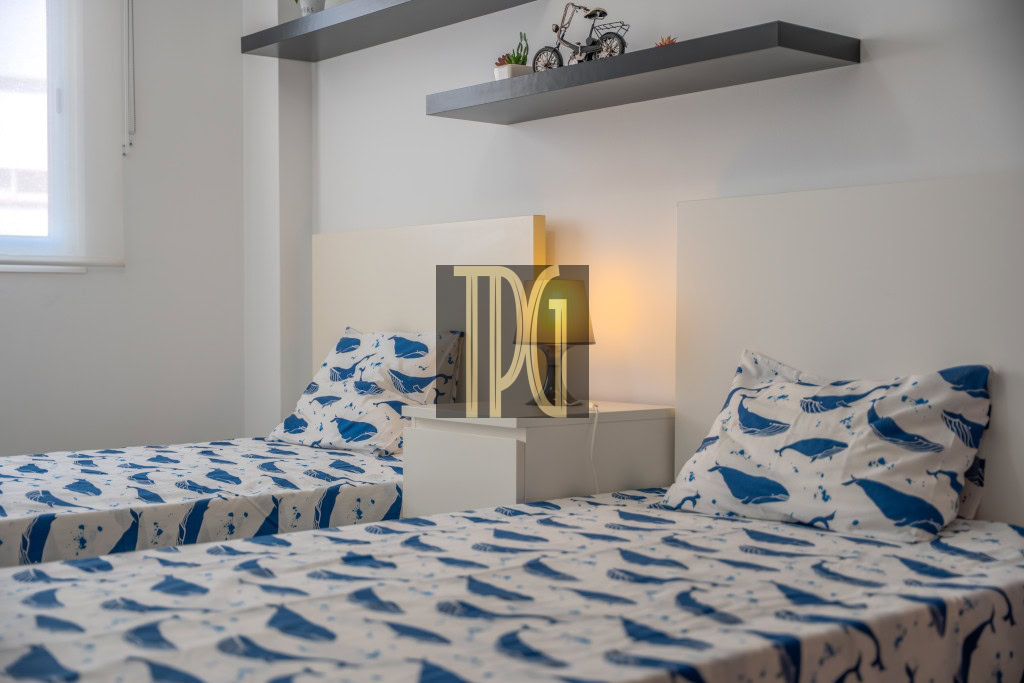 Twin bedroom with two single beds covered in blue whale-pattern bedding, a white nightstand between them, and a lit bedside lamp.