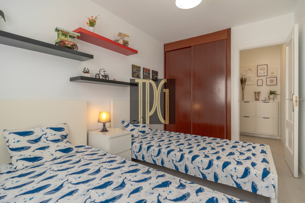 Twin bedroom with white headboards, two single beds in whale-pattern blue and white bedding, a nightstand lamp between them, and wall shelves with toys and decor.