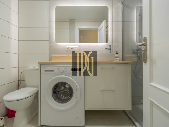 Modern bathroom with a front-loading washing machine under a wooden countertop, a sink and glass-door shower; toilet on the left.
