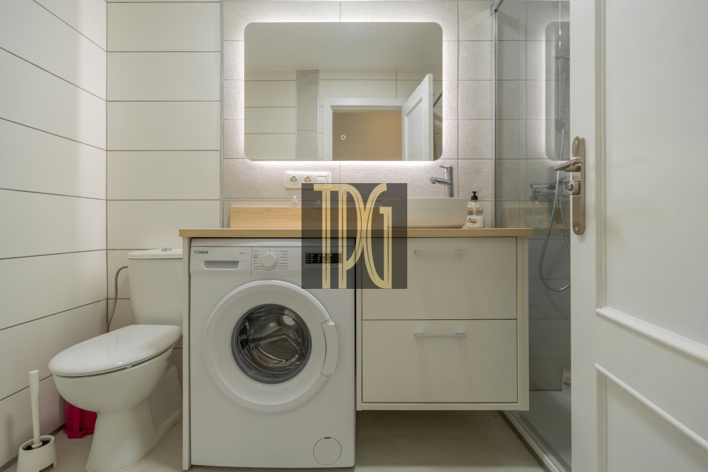 Modern bathroom with a front-loading washing machine under a wooden countertop, a sink and glass-door shower; toilet on the left.