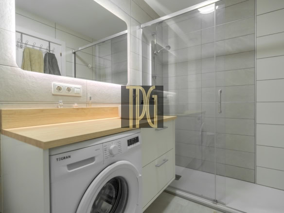 Modern bathroom with a front-loading washing machine under a wooden countertop, white cabinets, large mirror, and a glass-enclosed shower.