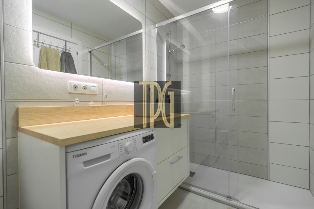 Modern bathroom with a front-loading washing machine under a wooden countertop, white cabinets, large mirror, and a glass-enclosed shower.
