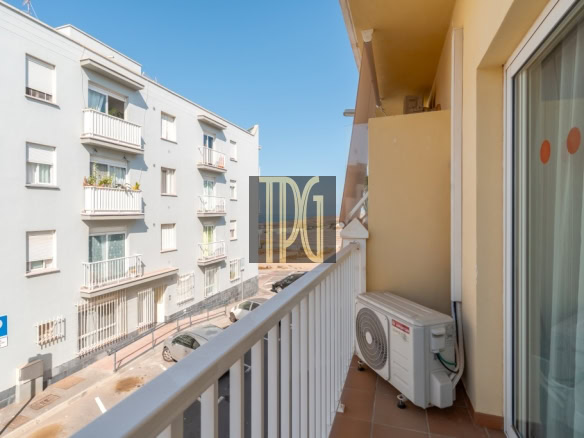 Balcony with an outdoor air conditioner overlooking a sunny urban street lined with white apartment buildings and parked cars.