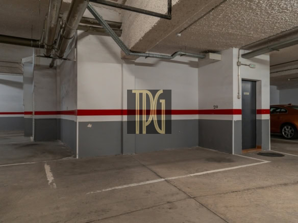 Indoor parking garage with a red stripe along gray-and-white walls, concrete ceiling, and exposed pipes; a gray door labeled 20 and an orange car to the right; centered TDG logo overlay.