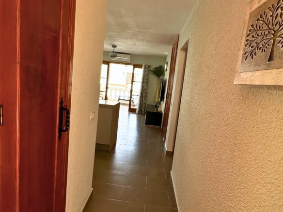 Narrow hallway with brown tile floor and a wooden door on the left, wall art on the right, leading to a sunlit living area with glass doors at the end.