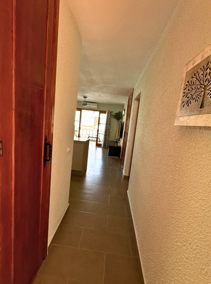 Narrow hallway with brown tile floor and a wooden door on the left, wall art on the right, leading to a sunlit living area with glass doors at the end.