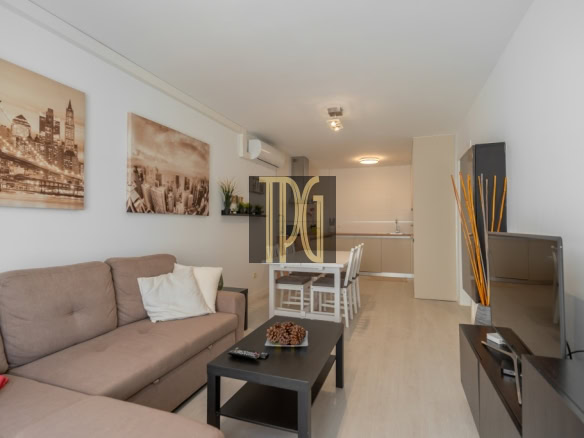 Open-plan living room with a beige sofa, cityscape art on the wall, and a dining table near a compact kitchen area.