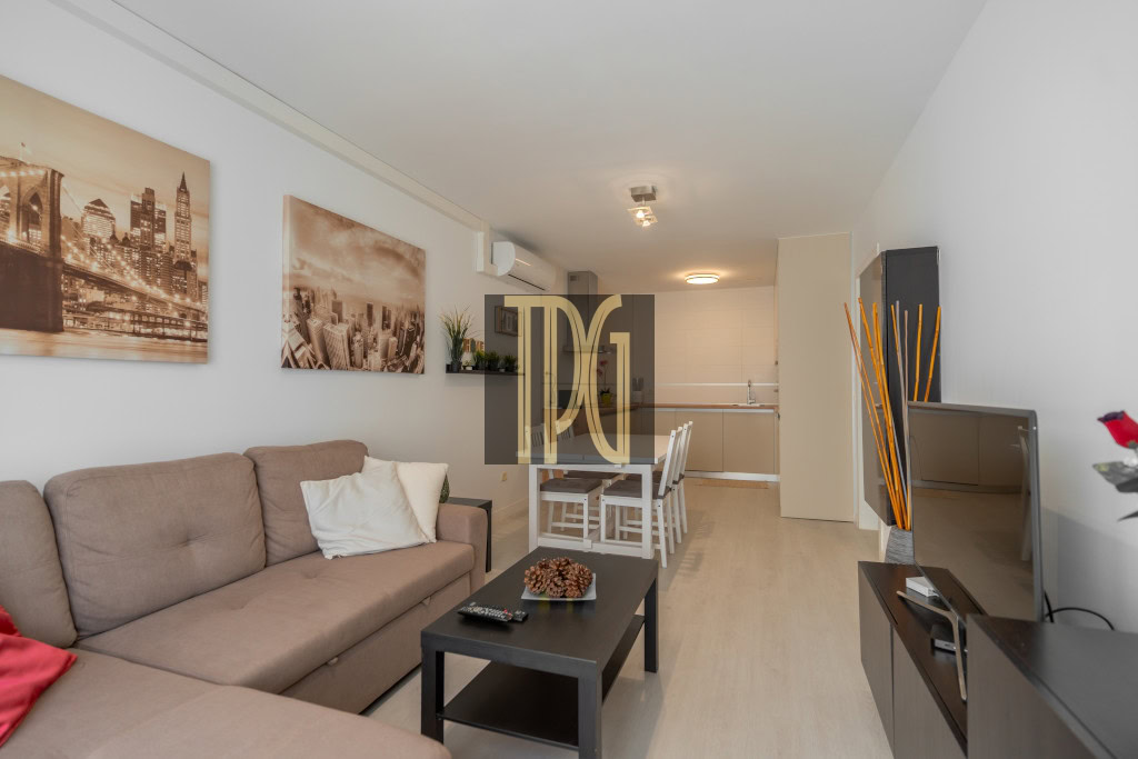 Open-plan living room with a beige sofa, cityscape art on the wall, and a dining table near a compact kitchen area.
