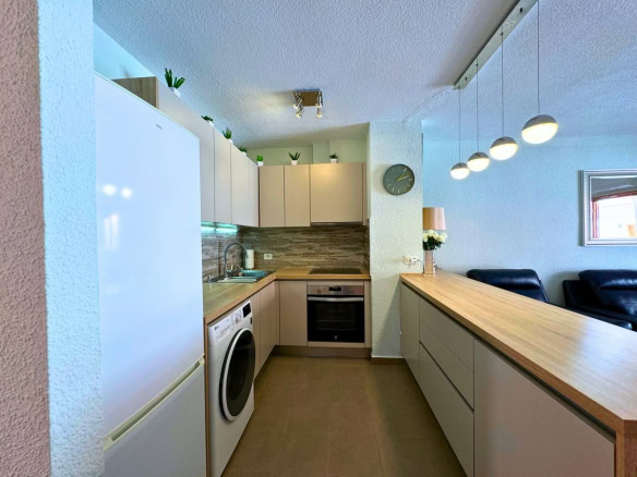 Open-plan kitchen with white and beige cabinets, wooden countertops, island bar, and built-in appliances (fridge, washing machine, oven). Plants on top cabinets and pendant lights above the bar are visible, with a living area seating in the background.