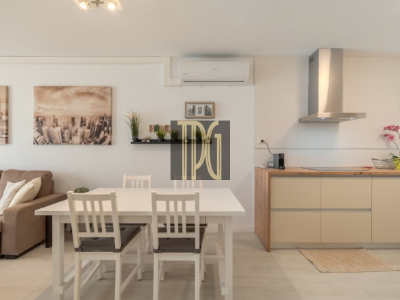 Open-concept living area with a white dining table and four chairs, beige sofa, cityscape wall art, and a kitchen with a wooden countertop and range hood down the wall.