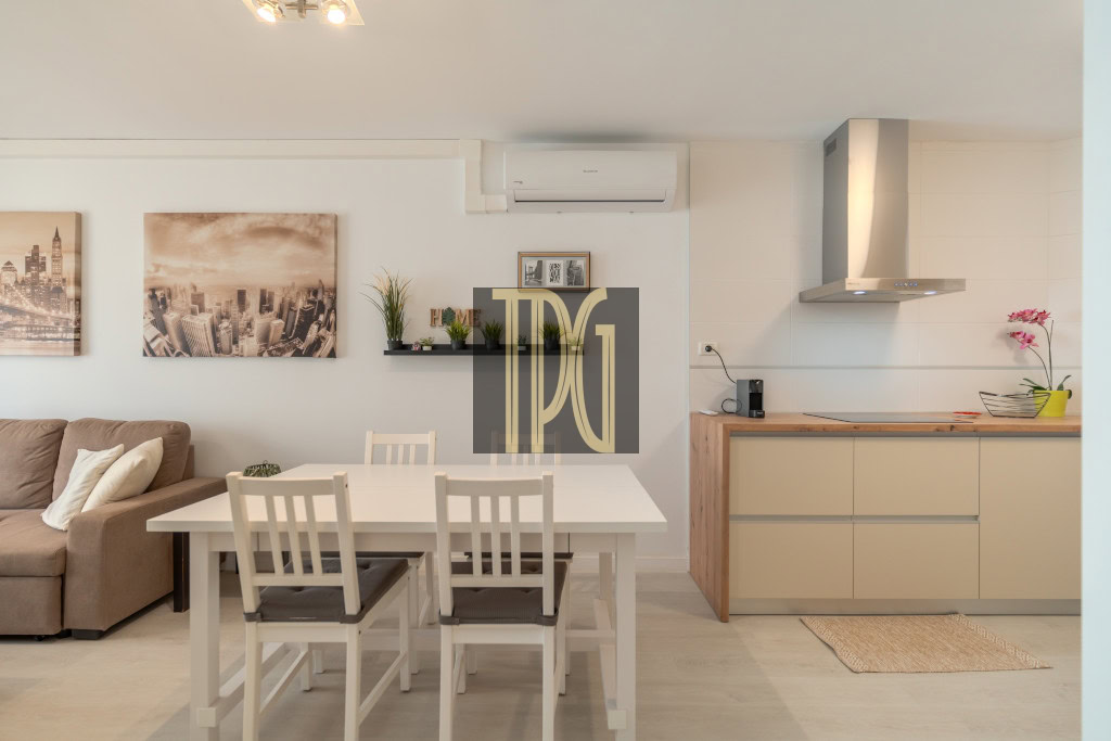 Open-concept living area with a white dining table and four chairs, beige sofa, cityscape wall art, and a kitchen with a wooden countertop and range hood down the wall.