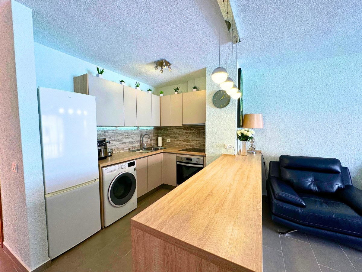 Open-plan kitchen with white cabinets, beige backsplash, wooden island, and a black leather sofa in the living area.