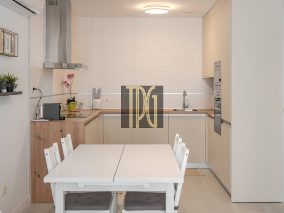 Bright white kitchen with a white dining table and chairs in the foreground, wooden counter and built‑in appliances in the back.