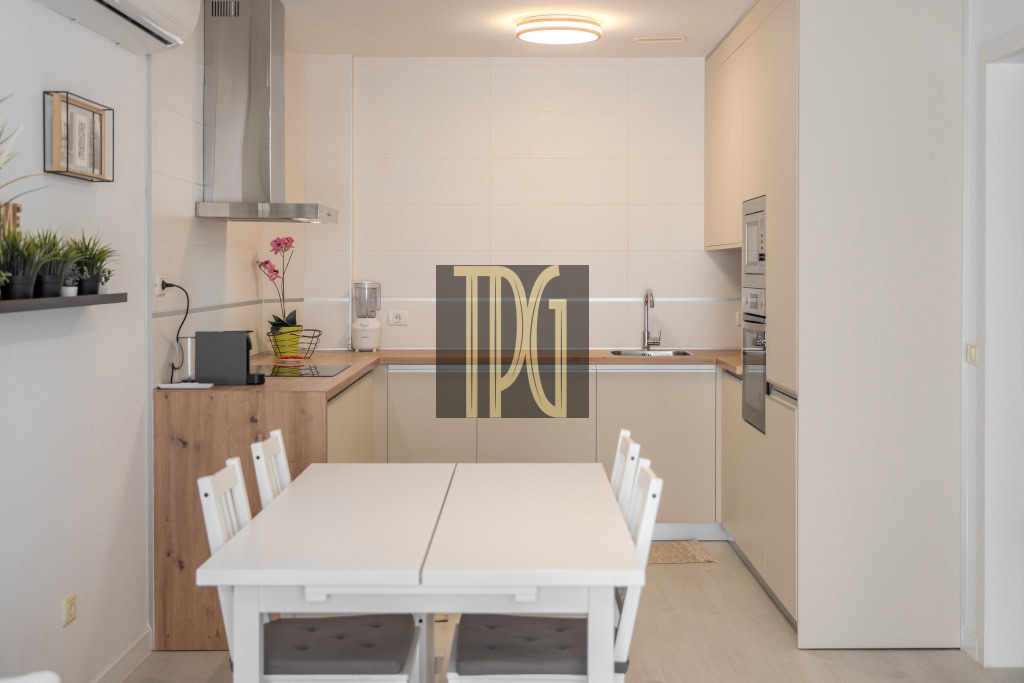 Bright white kitchen with a white dining table and chairs in the foreground, wooden counter and built‑in appliances in the back.