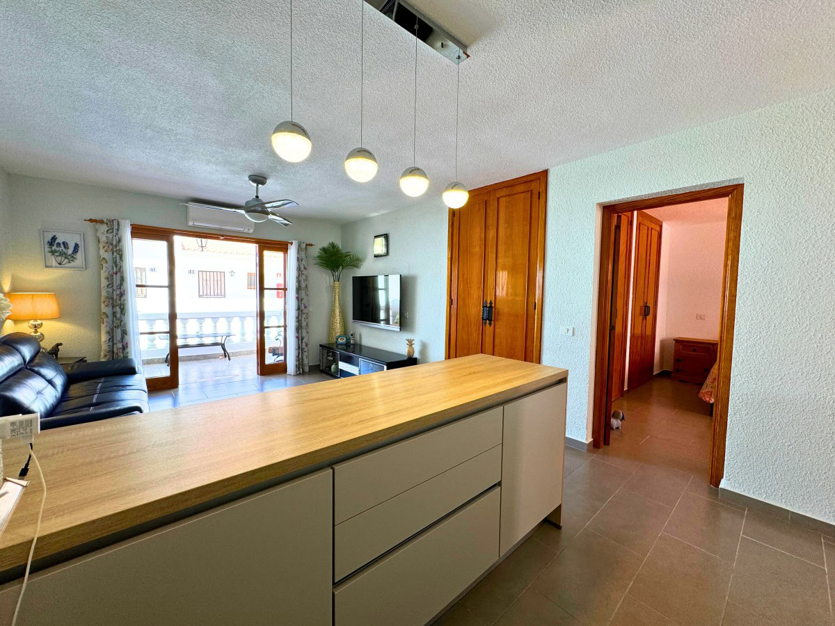 Open-plan living room with a wooden kitchen island, gray cabinetry, a black leather sofa, TV, and glass doors opening to a balcony.” (informative)